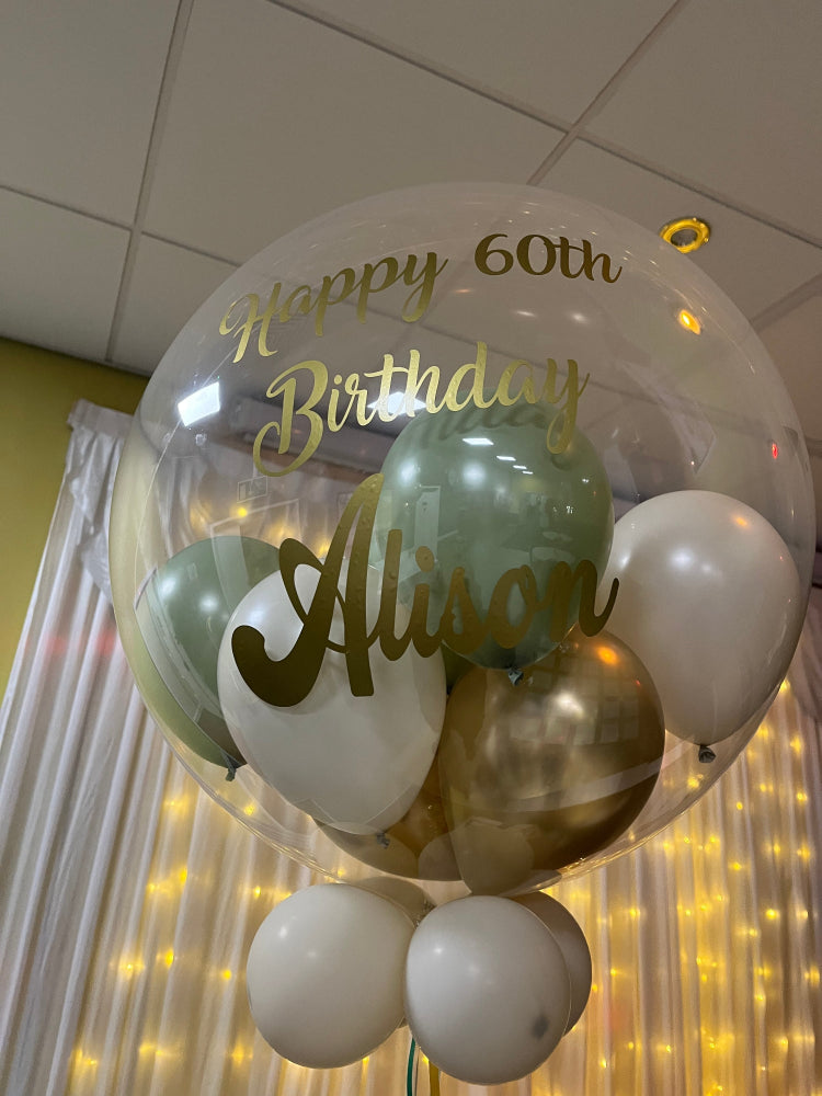 Helium inflated 24 inch personalised bubble balloon, this example shows metallic gold, white sand, eucalyptus mini balloons with gold personalisation.