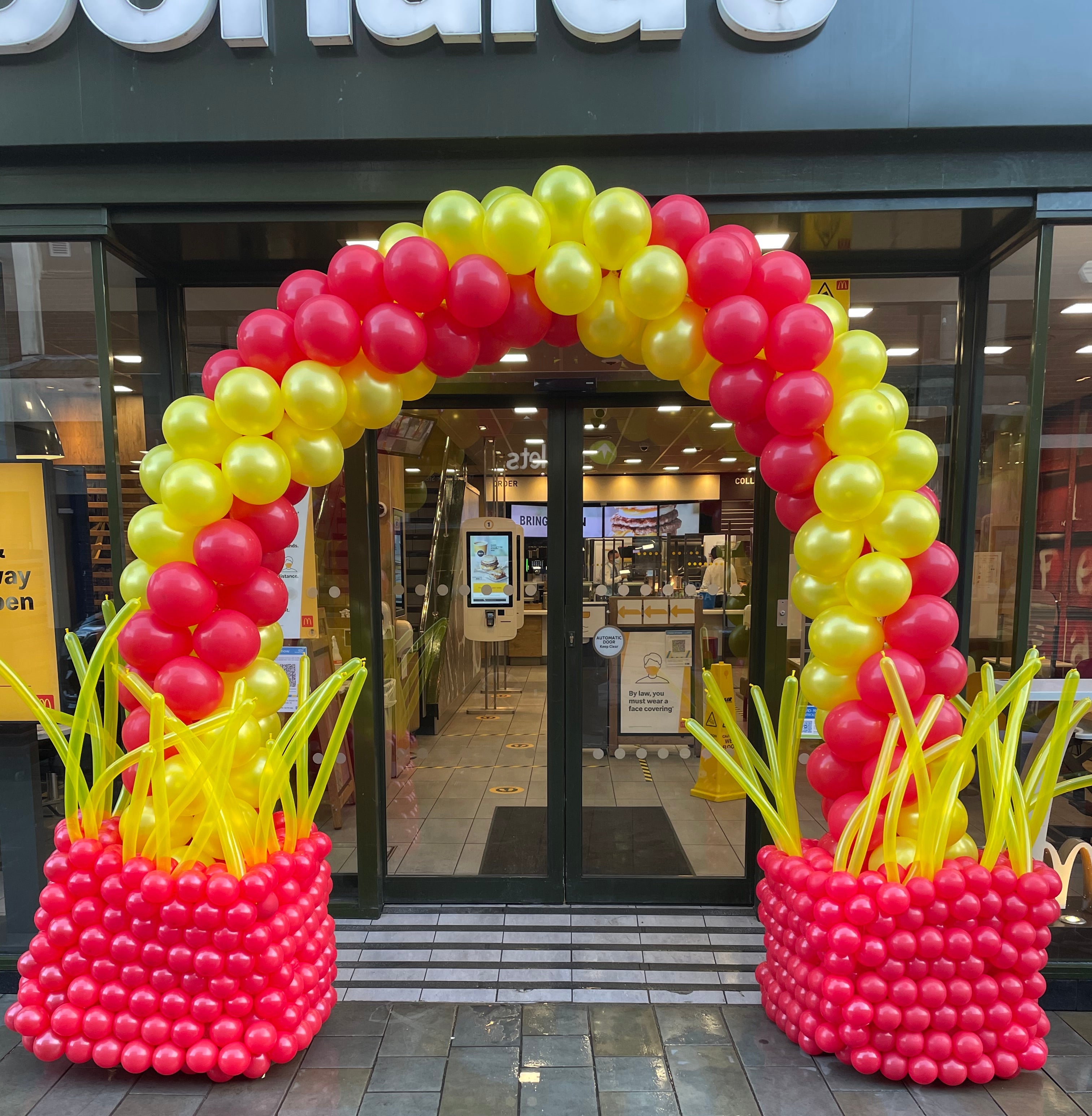 Balloon arch with red, yellow, and pink balloons in front of a building entrance.